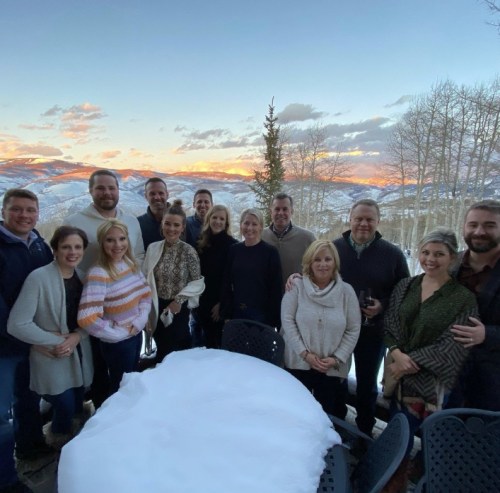 A group of people poses together outdoors with a snowy landscape and sunset in the background.
