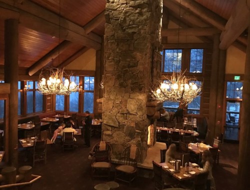 Cozy restaurant interior with wooden beams, stone fireplace, and chandeliers, featuring tables and diners in warm lighting.