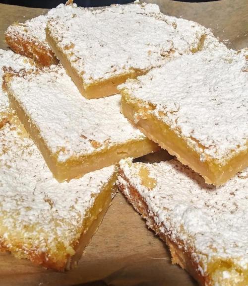 A close-up of lemon bars dusted with powdered sugar, arranged on a brown paper surface.