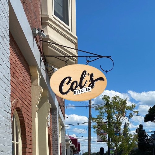 Sign for "Col's Kitchen" hanging from a building, with a blue sky and clouds in the background.