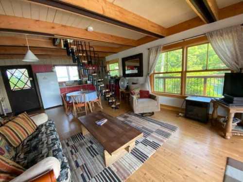 Cozy living room with wooden beams, a staircase, and large windows overlooking greenery. Features a sofa, table, and TV.
