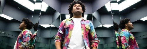 A person with curly hair stands in a mirrored room, wearing a colorful tie-dye shirt and looking upward.