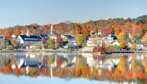 A serene lakeside view of a town with colorful autumn foliage reflecting on the water.