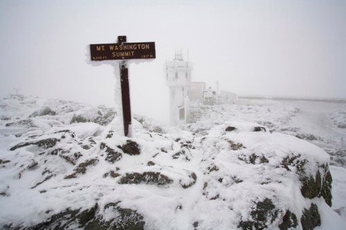 Snow-covered rocks and a sign reading "Mt. Washington Summit" in a foggy, wintry landscape.