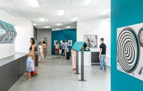 A modern art gallery with visitors exploring exhibits and interactive displays on teal and gray walls.