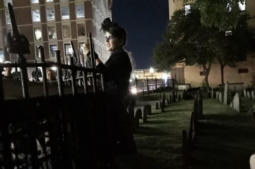 A person in dark clothing stands by a wrought iron fence at night, with a cemetery and city lights in the background.