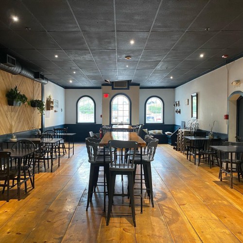 A spacious restaurant interior with wooden floors, black chairs, and large windows, featuring a cozy, inviting atmosphere.