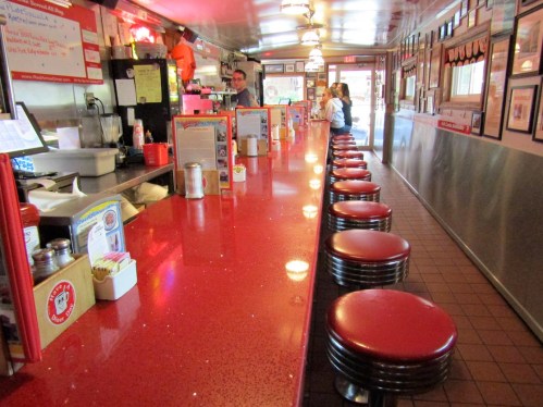 A retro diner interior with a shiny red counter, chrome stools, and a few patrons seated at the bar.