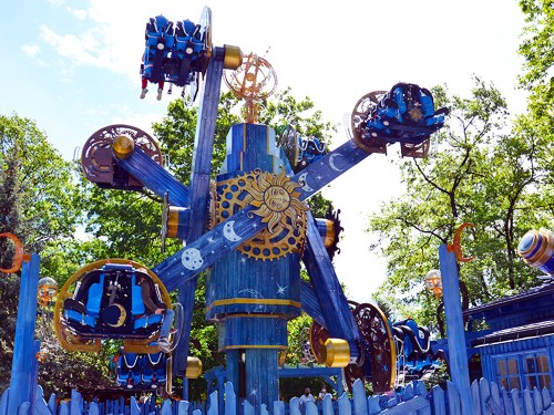 A colorful amusement park ride with a sun motif, featuring multiple seats and a whimsical design, surrounded by trees.