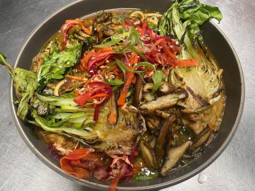 A bowl of vibrant noodle soup topped with greens, mushrooms, and colorful pickled vegetables.