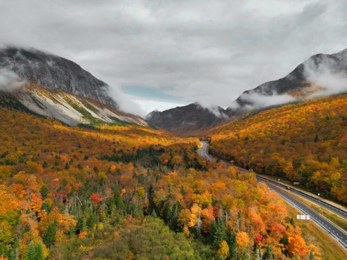 Aerial view of a winding road through vibrant autumn foliage and mountains under a cloudy sky.