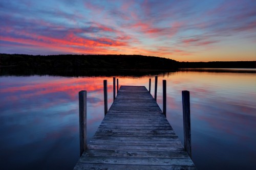 A wooden dock extends into a calm lake, reflecting a vibrant sunset with pink and orange hues in the sky.