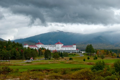 A grand hotel with red roofs sits amidst green trees and mountains under a cloudy sky.