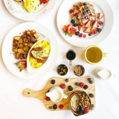 A brunch spread featuring eggs, potatoes, waffles with berries, and a wooden board with oatmeal and toppings.