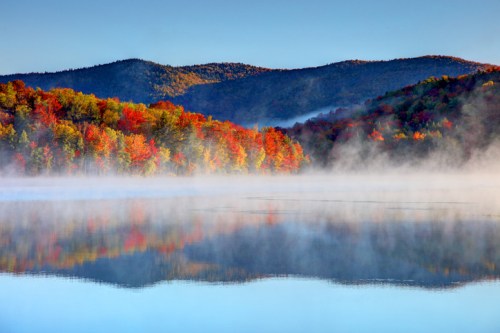 A serene lake surrounded by vibrant autumn foliage and misty mountains under a clear blue sky.