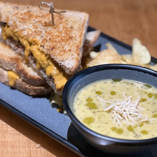 A grilled cheese sandwich with melted cheese, served with a bowl of creamy soup and a side of potato chips.