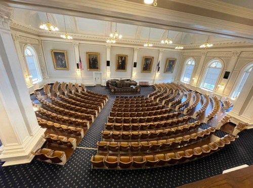 Interior view of a legislative chamber with rows of empty seats and a speaker's podium, featuring large windows and portraits.