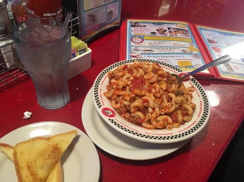 A plate of pasta with vegetables and pepperoni, a glass of water, and a slice of toast on a red table.