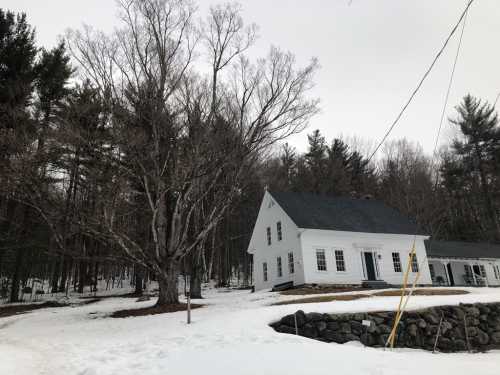 A white house with a dark roof surrounded by snow and bare trees, set against a backdrop of a forest.