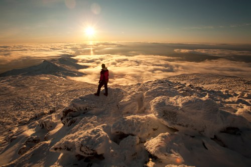 A person stands on a snowy mountain peak, overlooking clouds and a sunset in the background.