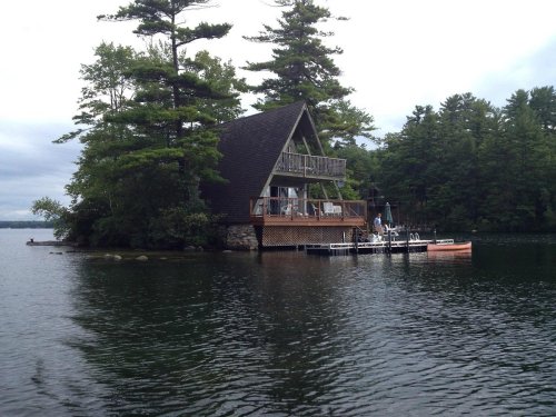 A lakeside cabin surrounded by trees, with a dock and small boat in the foreground, under a cloudy sky.