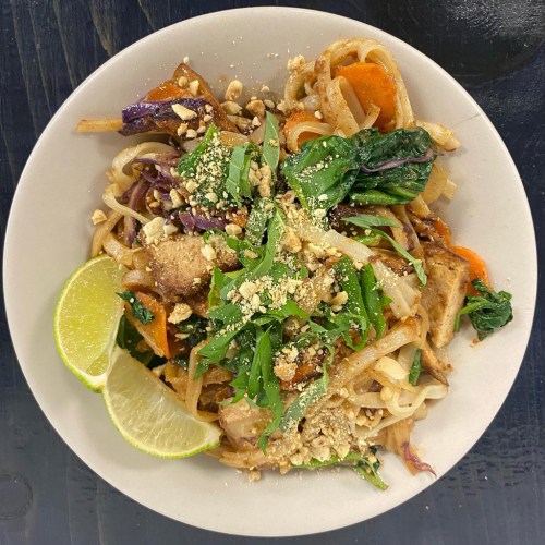 A plate of stir-fried noodles with vegetables, topped with crushed peanuts and lime wedges.