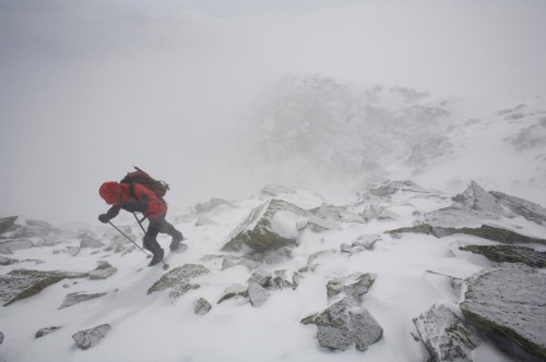 A climber in a red jacket ascends a rocky, snow-covered mountain in foggy, harsh weather conditions.