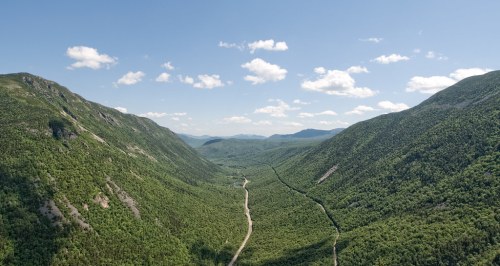 Aerial view of a lush green valley surrounded by mountains under a clear blue sky with scattered clouds.