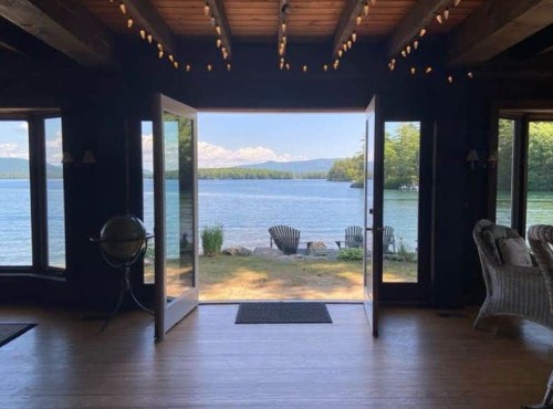 A serene lake view from a cozy interior with open doors, featuring chairs and mountains in the background.