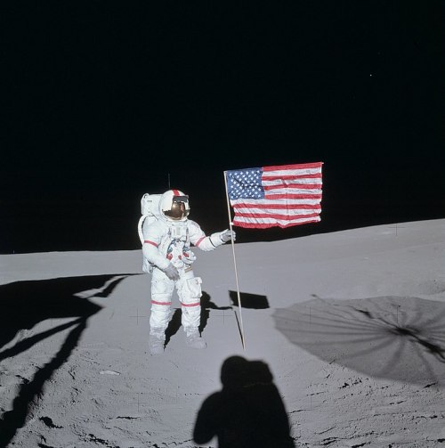 An astronaut stands on the moon, holding an American flag, with a shadow cast on the lunar surface.