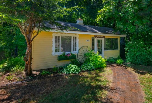 A charming yellow cottage with a brick path, surrounded by greenery and a decorative wheel by the window.