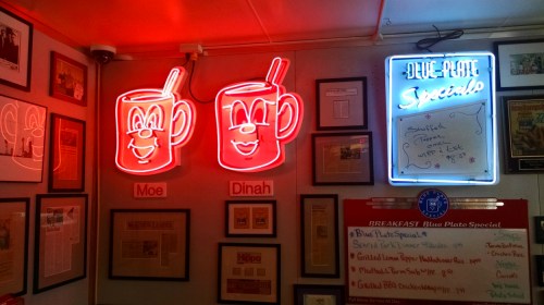 Neon signs of two smiling mugs named Moe and Dinah, surrounded by framed articles and a special menu board.