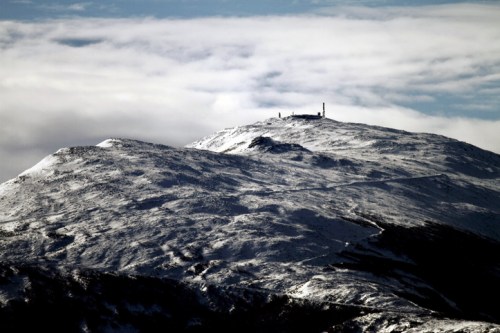 Snow-covered mountain peak under a cloudy sky, with structures visible at the summit.