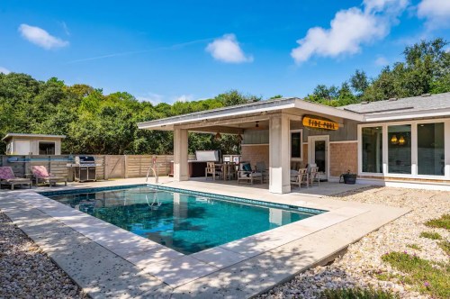 Modern backyard with a pool, patio seating, and lush greenery under a clear blue sky.