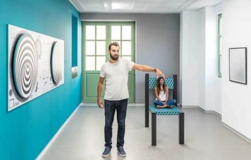 A man stands beside a large chair, while a woman sits inside it, in a modern art gallery with colorful walls.