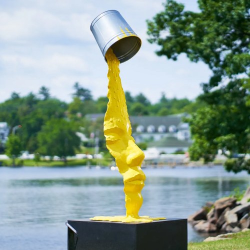 A yellow paint can spills paint, forming a face sculpture, set against a scenic lakeside background.