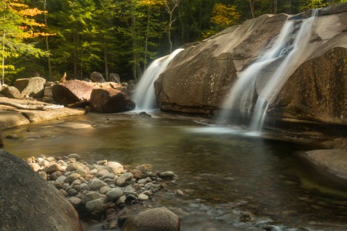 A serene waterfall cascades over rocks into a calm pool, surrounded by lush green trees and autumn foliage.