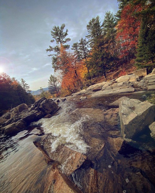 A serene river flows over rocks, surrounded by autumn trees and a clear sky, creating a peaceful natural scene.