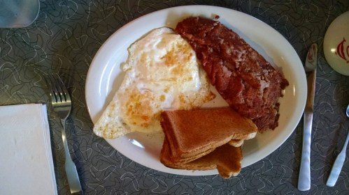 A plate with a fried egg, a slice of meatloaf, and two pieces of toasted bread on a patterned table.