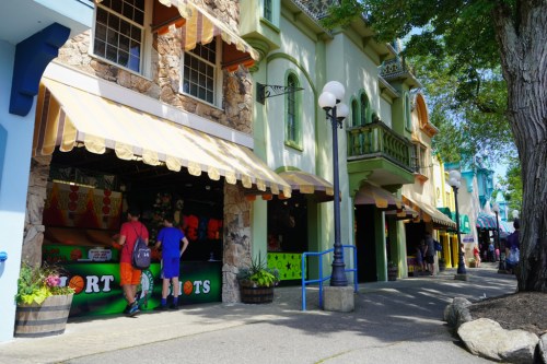 Colorful storefronts line a pathway, with people walking and enjoying a vibrant outdoor market atmosphere.