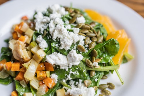 A vibrant salad with spinach, roasted vegetables, pumpkin seeds, and crumbled feta cheese on a white plate.