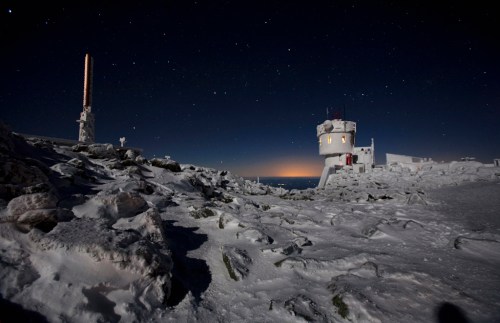 A snowy landscape at night featuring a tower and a building under a starry sky, with a distant glow on the horizon.