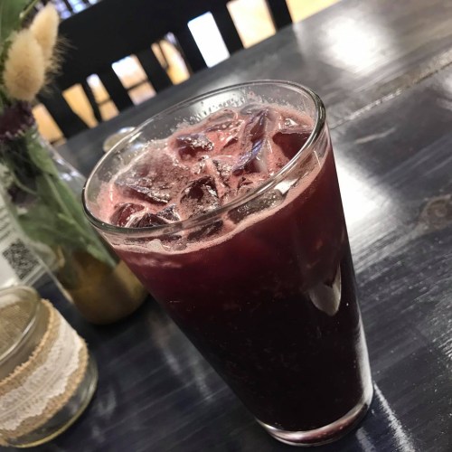 A glass of dark purple beverage with ice, sitting on a wooden table next to a decorative jar and a flower vase.