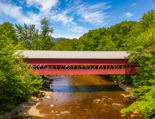 A red covered bridge spans a clear river, surrounded by lush green trees and a blue sky with scattered clouds.