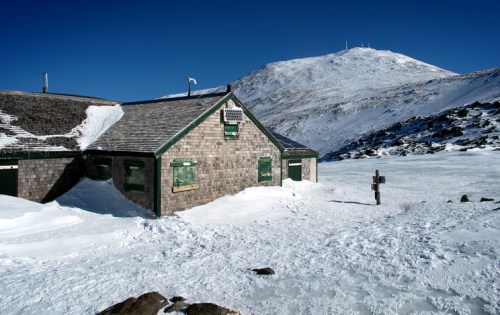 A snow-covered building with green windows, surrounded by a winter landscape and a mountain in the background.