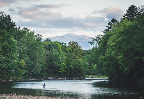 A person wades in a calm river surrounded by lush trees, with mountains in the background under a cloudy sky.