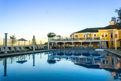 A serene pool area with lounge chairs, surrounded by a yellow building and reflecting the clear blue sky at sunset.