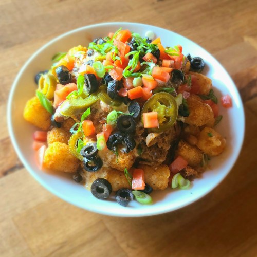 A bowl of loaded tater tots topped with jalapeños, olives, tomatoes, and green onions.