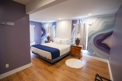 A modern bedroom with a large bed, wooden nightstands, and colorful abstract wall art. Natural light from windows.