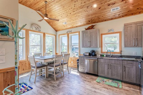 Bright kitchen with wooden ceiling, large windows, a dining table, and modern appliances. Coastal decor accents.